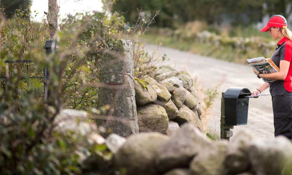 A woman in a Posten uniform who putting mail into a black mailbox