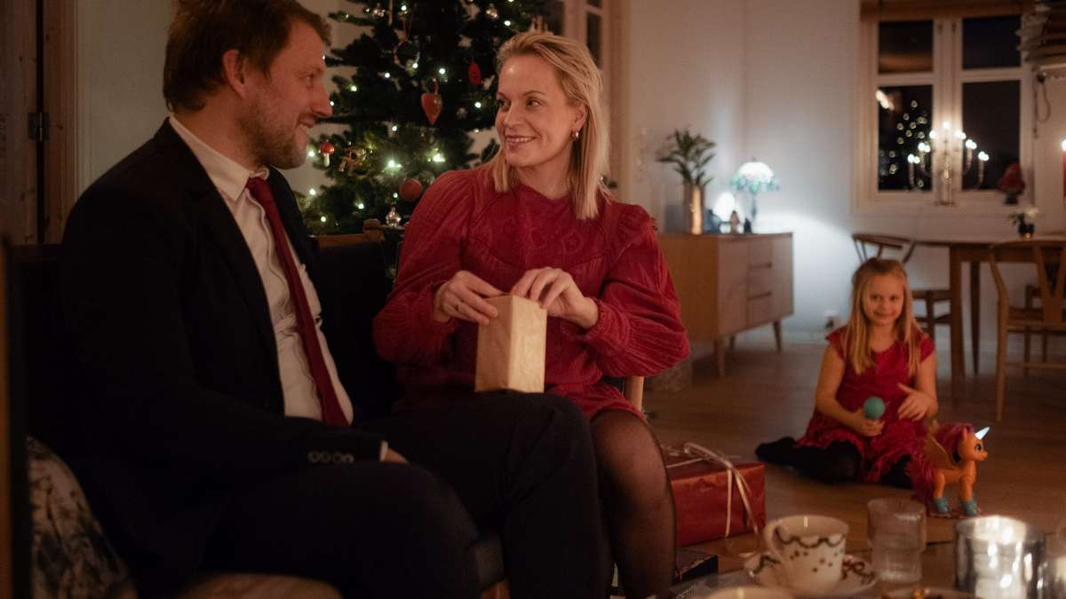 kvinne åpner julegave foran juletre, sammen med mann og barn.