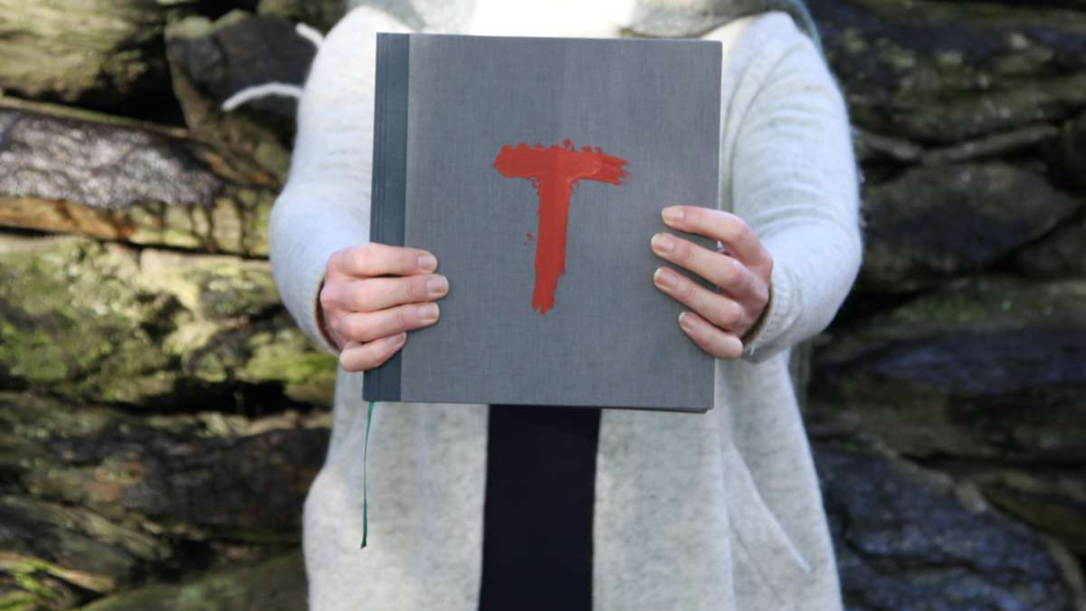 En person som holder frem en grå bok som har en rød T på forsiden